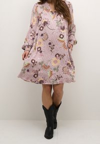 Vestido floral de tela rosa claro con mangas largas, escote en V y bolsillos. Presenta patrones de flores multicolores y un ajuste relajado. Combinado con botas negras.