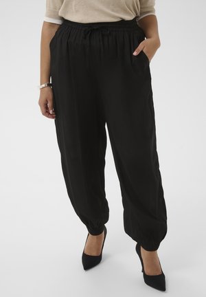 Žena má na sebe voľné čierne nohavice s elastickými manžetami a vreckami, v kombinácii s béžovým topom a čiernymi špicatými vysokými podpätkami.