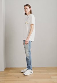 T-shirt de algodón blanco con diseño gráfico, combinado con jeans rotos de color azul claro y zapatillas blancas sobre un suelo de madera. Fondo neutro.