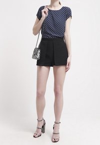 Marineblaues Polka-Dot-T-Shirt, schwarze hoch taillierte Shorts, silberne Umhängetasche und Sandalen mit Blockabsatz. Glatter Stoff, taillierte Passform.