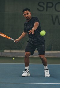 Jugador de tenis con una camiseta negra de manga corta y pantalones cortos, usando una raqueta gris. Zapatillas blancas notables con acentos en negro y verde.