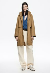 Cappotto impermeabile marrone con cappuccio, grandi tasche e chiusure a scatto, indossato sopra una camicia blu e pantaloni beige. Sneakers grigie completano il look.