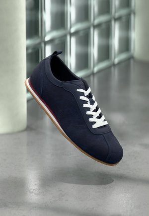 Marineblå afslappet sneaker med hvide snørebånd, gummisål og rød stribe, svævende over en blank betongulv nær glasblokke.