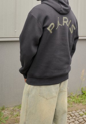 Personne portant un sweat à capuche foncé avec « PARIS » et un logo silhouette dans le dos, associé à un jean large de couleur claire, debout à l'extérieur.