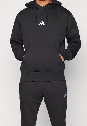 Person trägt schwarzen Adidas-Hoodie und passende schwarze Adidas-Sweatpants mit weißen Logos, Hände in der vorderen Tasche des Hoodies.