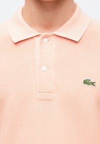 Ανοιχτού ροζ πόλο μπλούζα με κολάρο, διαθέτει τρία κουμπιά και πράσινο κεντημένο λογότυπο κροκοδείλου στην αριστερή πλευρά. Υφασμα με υφή.