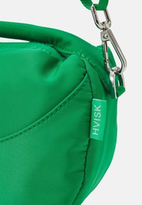 Angolo di una borsa di stoffa verde brillante con etichetta HVISK e un gancio in metallo argento che fissa una tracolla verde coordinata.
