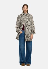 Beżowy płaszcz w ciemne plamy, z długimi rękawami, ko łnierzem i otwarciem z przodu; zestawiony z oversize'owymi niebieskimi dżinsami i brązowymi butami.