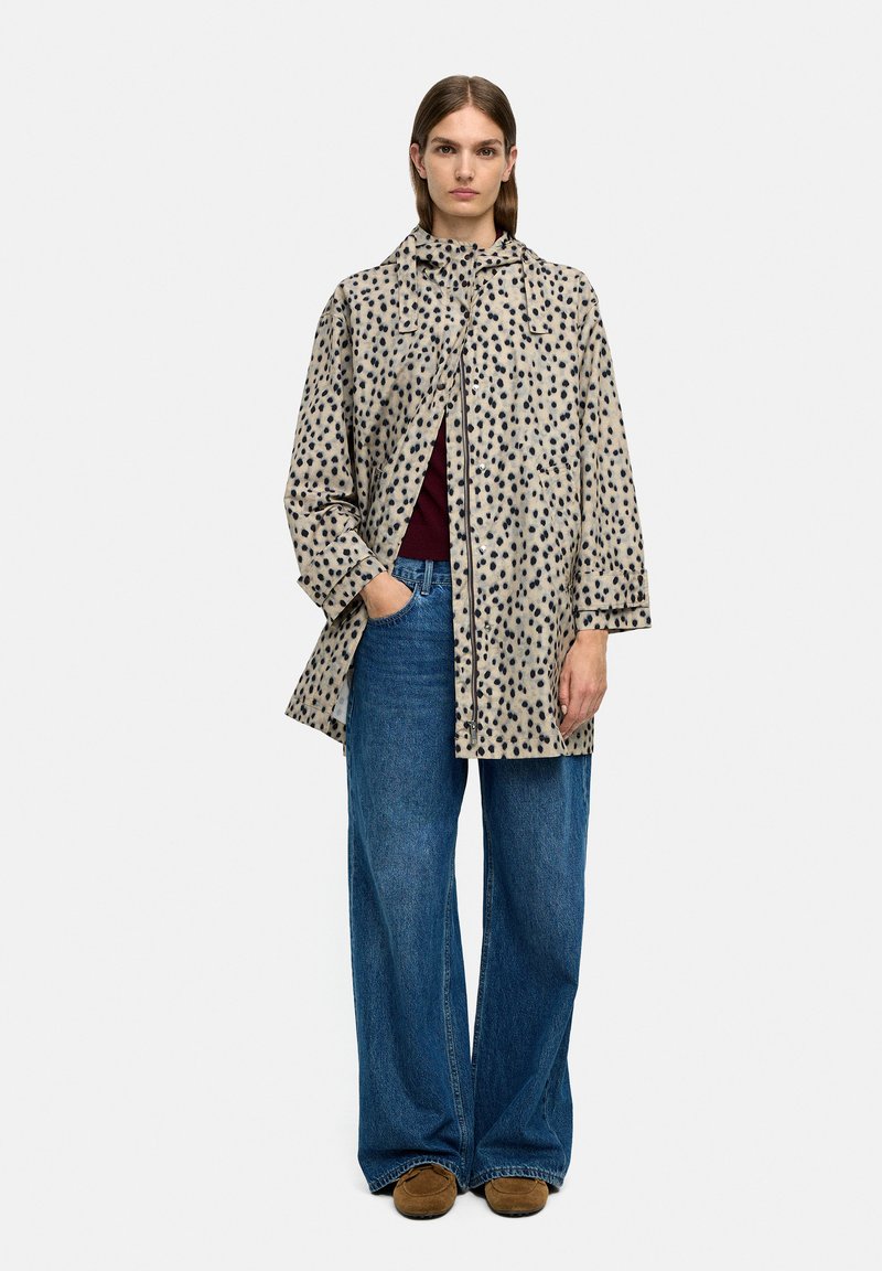 Beżowy płaszcz w ciemne plamy, z długimi rękawami, kołnierzem i otwarciem z przodu; zestawiony z oversize'owymi niebieskimi dżinsami i brązowymi butami.