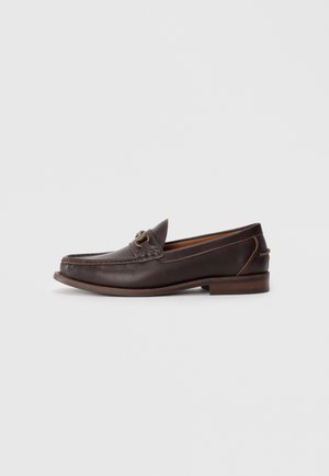 Mocasin de cuero marrón oscuro con detalle de hebilla metálica, tacón plano y costura estilo mocasín sobre un fondo claro.