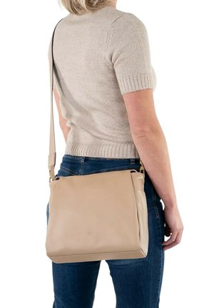 Donna che indossa un maglione beige a maniche corte e jeans, porta una borsa a tracolla in pelle beige con tracolla regolabile sulla spalla.