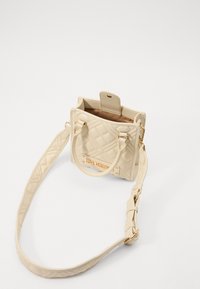 Beżowa torebka pikowana ze złotymi akcentami, górnymi uchwytami, regulowanym paskiem oraz emblematem na przodzie. Otwierane wnętrze ujawnia beżowe wykończenie.