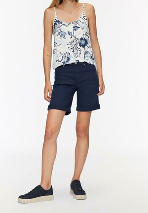 T-shirt sans manches à motif floral bleu associé à un short marine à revers et des baskets sombres avec des semelles texturées. Design simple et décontracté.