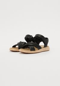 Sandale noire avec des bandes croisées, fabriquée en matériau doux. Dispose d'une semelle texturée couleur tan et d'une bride de cheville rembourrée pour plus de confort.