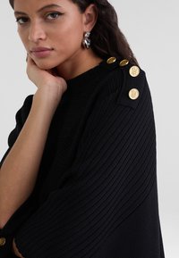 Schwarzer ribbeliger Pullover mit Rundhalsausschnitt und dekorativen goldenen Knöpfen an der Schulter. Weiche Textur mit lockerer Passform.