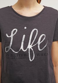 Mørk grå t-shirt med stort paliettekst "Life" og mindre trykt tekst nedenfor, der læser "er bedre når du griner."