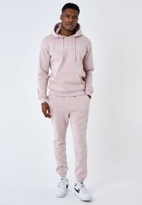 Conjunto de sudadera con capucha y pantalones jogger en suave tejido de color rosa claro, con un bolsillo frontal, cordones y un logo blanco. Lleva zapatillas blancas.