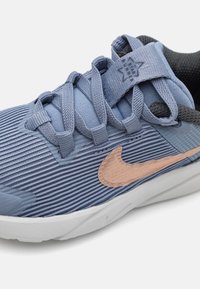 Chaussure de sport bleue avec tige texturée, tissu côtelé, accents gris et un logo Swoosh marron clair. Semelle blanche et zone de renforcement à l'avant.