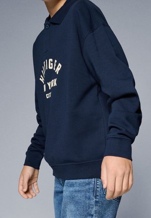 Barn iført navyblå langærmet skjorte med teksten "HILFIGER NEW YORK CITY", parret med lyseblå jeans, stående mod en neutral baggrund.