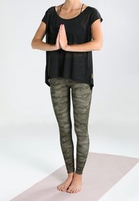 Top negro de manga corta con corte holgado, combinado con leggings de camuflaje verde oliva. Ambas prendas están hechas de materiales transpirables. Pose de yoga sobre la esterilla.
