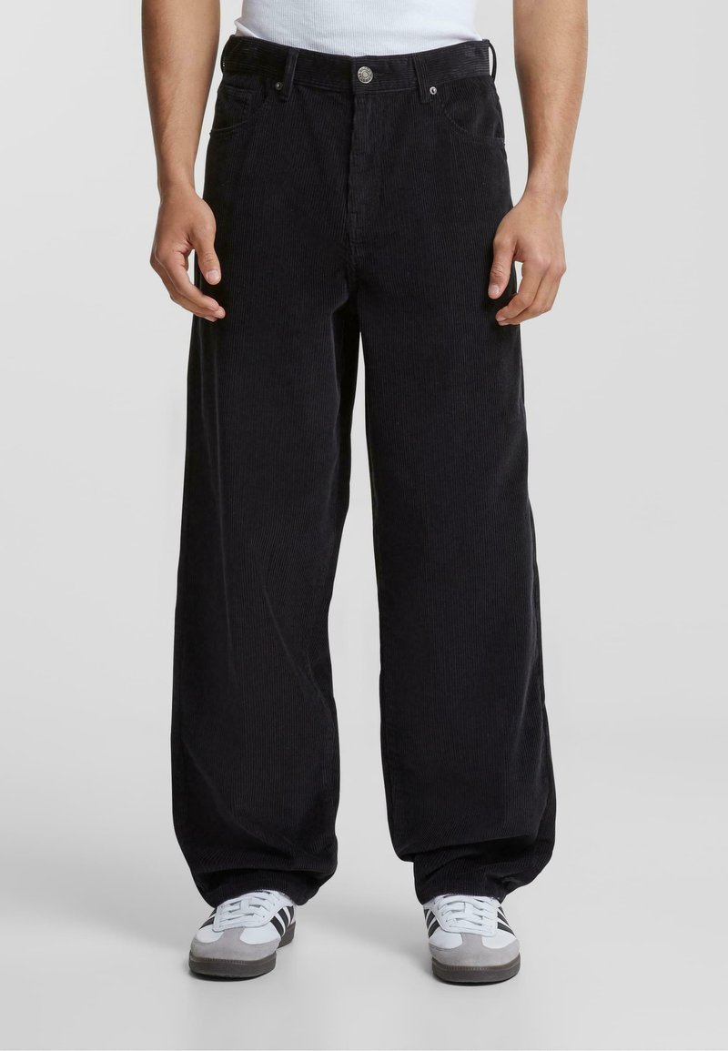 Pantaloni a gamba larga in corduroy nero con righe verticali, dotati di tasche frontali e chiusura con bottone. Abbinati a sneakers grigie e bianche.