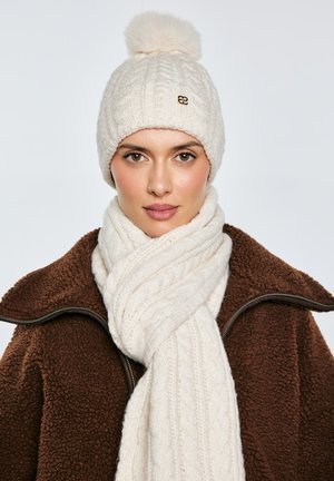 Moteris dėvi kreminės spalvos megztą kepurę su bumbulu ir derantį šaliką, rudą tekstūruotą striukę su užtrauktuku, atsisukusi į priekį priešais vientisą foną.