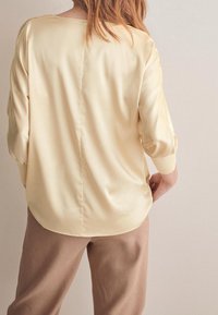 Blouse jaune en satin avec un ourlet arrondi et des manches trois quarts, associée à un pantalon beige ample, vue de dos.