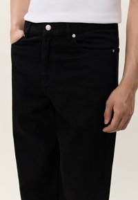 Κοντινό πλάνο ατόμου που φοράει μαύρο τζιν και λευκό πουκάμισο, με το ένα χέρι στην τσέπη και το άλλο χαλαρά στο πλάι.