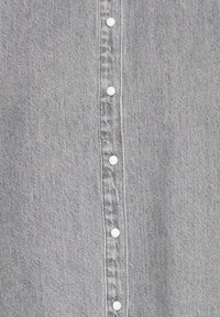 Chemise en denim gris avec une patte de boutonnage centrale ornée de boutons blancs, une texture subtile et des surpiqûres contrastantes le long des bords.