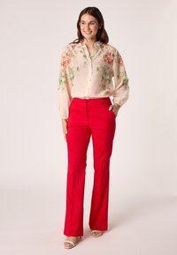 Bloes met bloemenprint in lichtroze, met knopen voor en lange mouwen, gecombineerd met felrode, uitlopende broeken en beige open teen hakken.