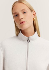 Weiße Zip-Jacke mit hohem Kragen, aus weichem Stoff. Verfügt über einen silbernen Reißverschluss und eine dezente Struktur, die eine moderne Ästhetik vermittelt.
