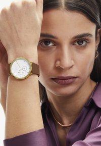 Ronde analoge horloge met een witte wijzerplaat, gouden kast, bruine siliconen band, met minimalistische markeringen en twee subdials voor de tijd.