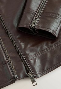 Ruskea nahkatakki metallisilla vetoketjuilla, lähikuva hihansuun ja etuvetoketjun yksityiskohdista sileällä tekstuurisella materiaalilla.