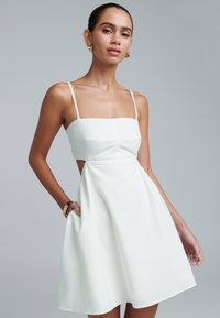 Hvid sommerkjole med firkantet halsudskæring, tynde stropper og cutout-detaljer i siden. Fremstillet af letvægtsstof med en udflaret silhuet.