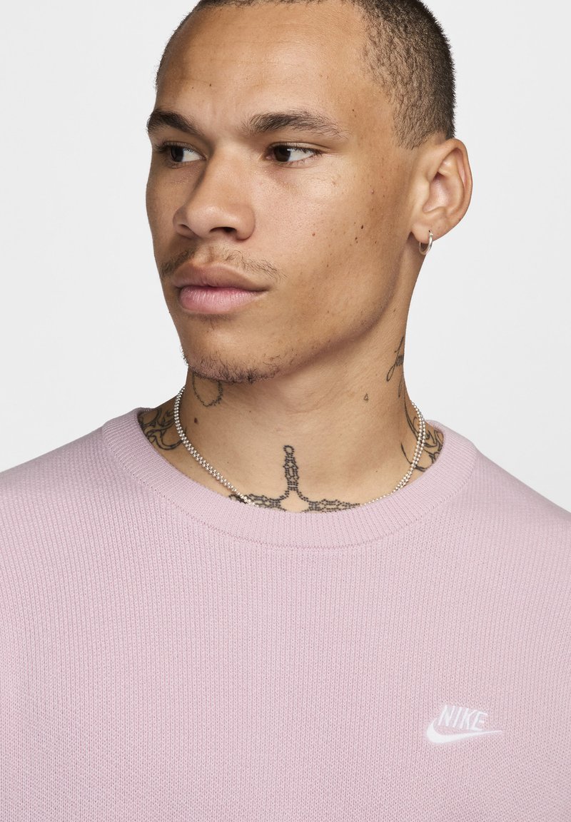 Fioletowy sweter z dzianiny z okrągłym dekoltem, z małym białym logo Nike na piersi. Modelka ma widoczne tatuaże i nosi warstwowe naszyjniki.
