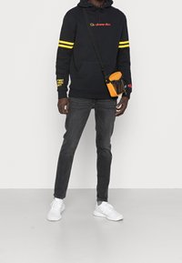 Felpa nera con strisce gialle, logo rosso, abbinata a jeans scuri e sneakers bianche. Include una piccola borsa a tracolla arancione.