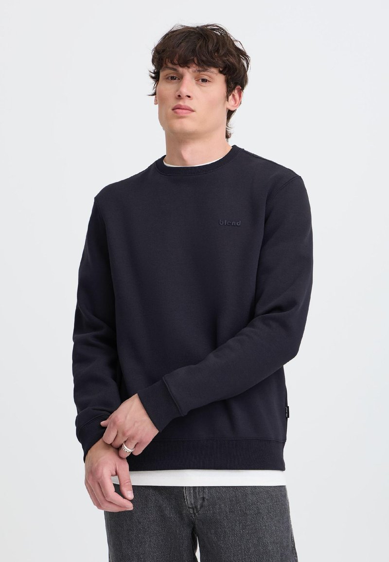 Ung mand med kort mørkt hår iført en sort crewneck sweatshirt, hvid undertrøje og mørke jeans mod en ensfarvet lys baggrund.