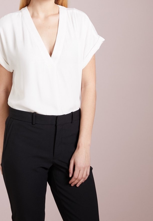 Kvinna klädd i en vit V-ringad blus med korta ärmar, instoppad i högmidjade svarta byxor framför en enkel beige bakgrund.