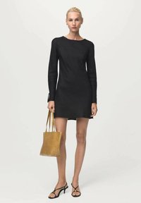 Una mujer está de pie vestida con un mini vestido negro de manga larga, tacones negros con tiras, y sostiene un bolso tote de cuero marrón frente a un fondo claro y liso.