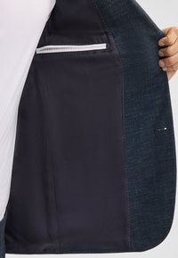 Blazer blu navy con un esterno texture e un livellamento blu navy liscio. Presenta una tasca interna con zip bianca e dettagli cuciti.