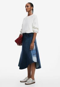 Jeanskjol med asymmetrisk nederkant, i olika blå nyanser och röd fickdetalj; matchad med en vit kabelstickad tröja och silverskor.