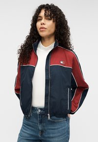 Μπουφάν με φερμουάρ σε ναυτικό μπλε και μπορντό χρώμα με λευκές λεπτομέρειες, διαθέτει ψηλό γιακά και λογότυπο στο στήθος, κατασκευασμένο από ελαφρύ ύφασμα.