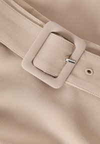 Beige riem gemaakt van gestructureerde stof, met een grote rechthoekige gesp en een zilveren pen, met zichtbare stikdetails.