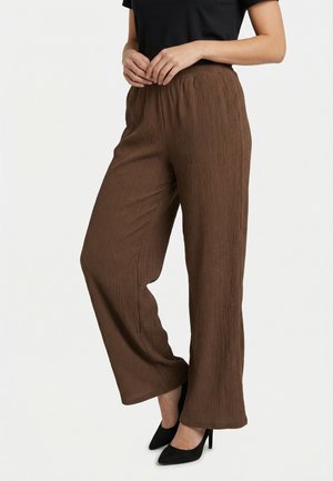 Mujer con pantalones marrones texturizados de talle alto y pierna ancha, combinados con zapatos de tacón alto negros y una camiseta negra metida por dentro.