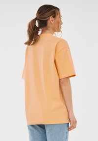 Overmaat t-shirt van zachte oranje katoen. Heeft korte mouwen, een ronde hals en een soepele textuur. G gedragen met lichtblauwe spijkerbroek.