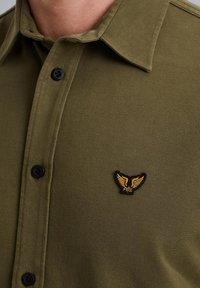 Camisa verde oliva con un tejido texturizado, con un cierre de botones negros y un pequeño logo de alas en negro y dorado en el pecho.