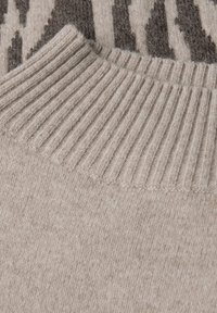 Beige gestrickter Pullover mit einem gerippten Ausschnitt und weicher Textur, im Hintergrund ist ein gemusterter Entwurf sichtbar.