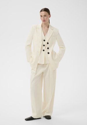 Femme portant un blazer croisé crème, un pantalon large assorti et des chaussures noires, debout les mains dans les poches sur fond blanc.