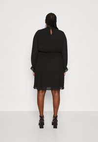 Robe noire plissée à manches longues, avec une découpe en clé dans le dos et une taille élastique. Accompagnée de sandales noires à talons carrés.