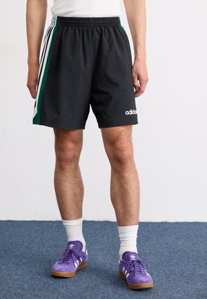 Shorts de sport noirs avec des accents latéraux rayés verts et blancs, ceinture élastique et poches. Le modèle porte des baskets violettes et des chaussettes blanches.